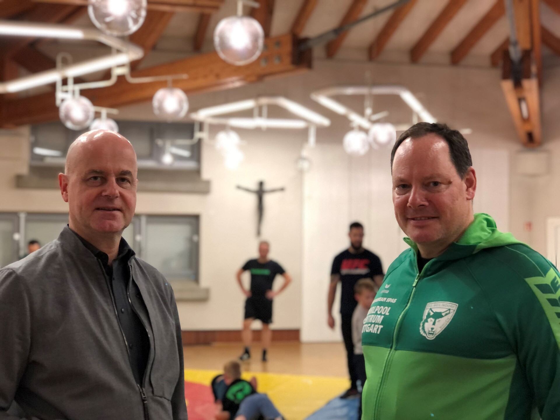 Pfarrer Matthias Hambücher (rechts im Foto) und Günter Buchholz (rechts), Jugendleiter bei der SG Weilimdorf stehen im Gemeindesaal der Gemeinde Salvator in Giebel. Im Hintergrund sind Jungen und Mädchen der Ringer beim Training zu sehen. 