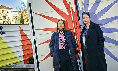 Zwei Frauen vor einem Container, der mit grafischen Elementen gestaltet ist.