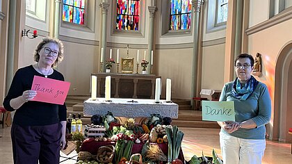 Zwei Frauen stehen neben Altar, in den Händen halten Sie Schilder mit dem Wort "Danke". Vor dem Altar sind Äpfel und andere Erntegaben schön aufgestellt