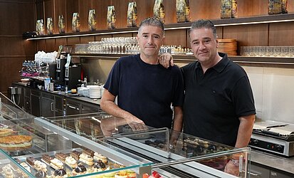 Die Brüder Vitsas Arm in Arm vor der Kuchentheke im Café Hegel im Haus der Katholischen Kirche