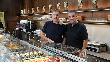 Die Brüder Nikolaos und Georgios Vitsas stehen nebeneinander vor der mit Kuchentheke